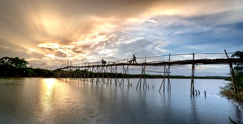 Free Workers crossing a bamboo bridge over a reflective river at sunset, under a dramatic sky. Stock Photo