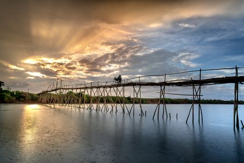 Free A serene view of a wooden bridge with a cyclist during sunset over calm water. Stock Photo