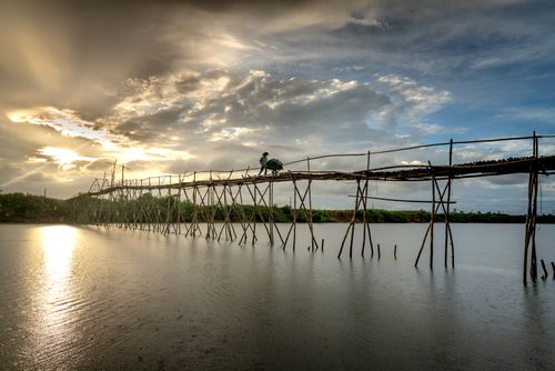 Free A narrow wooden bridge stretches over a tranquil lake at sunset with a person crossing, highlighting rural simplicity. Stock Photo