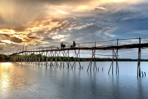 Free Two men crossing a bamboo bridge over a river at sunset with dramatic clouds. Stock Photo