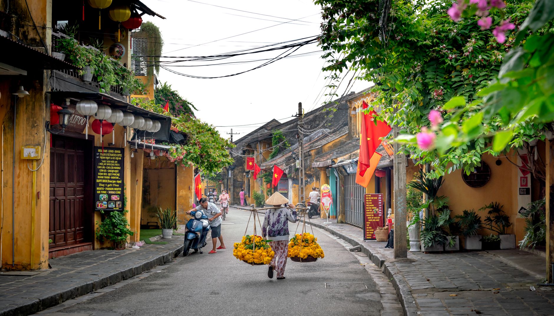 A vendor carrying flowers walks the streets of Hoi An, lined with Vietnamese flags and lanterns.