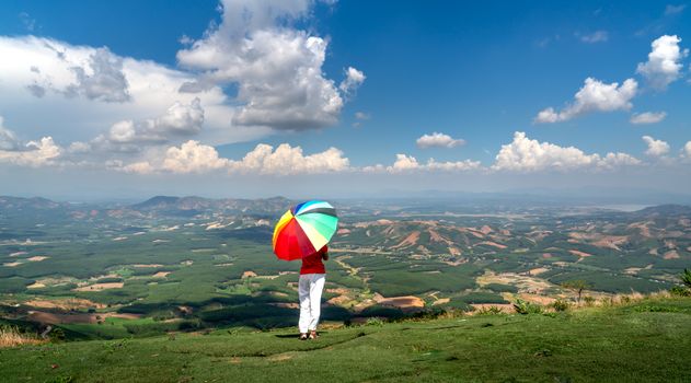 A person stands with a colorful umbrella overlooking a vast countryside landscape under a blue sky with clouds.