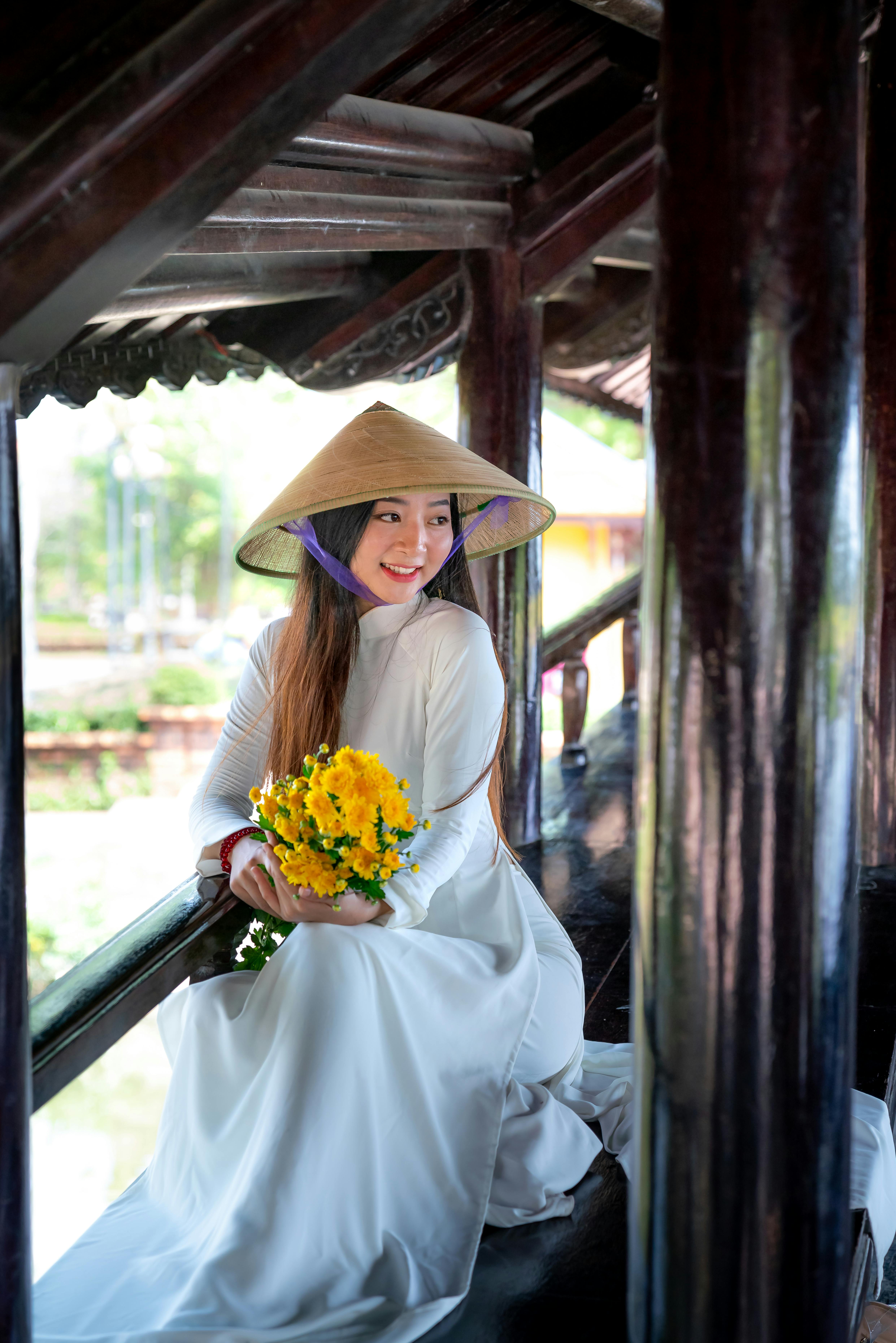 Beautiful woman in ao dai with a conical hat holding yellow flowers, seated outdoors.