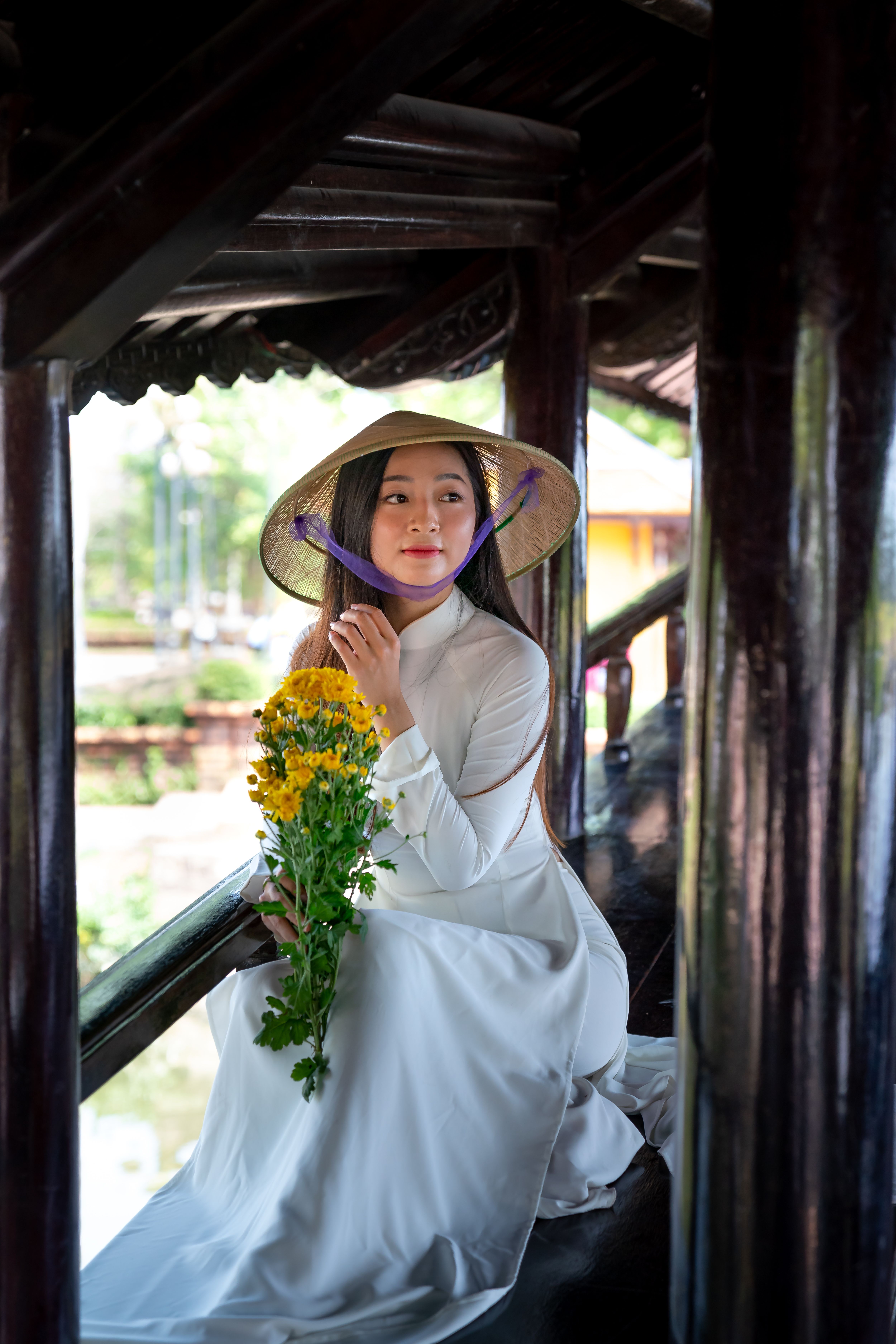 Asian woman in ao dai with conical hat holding yellow flowers indoors.