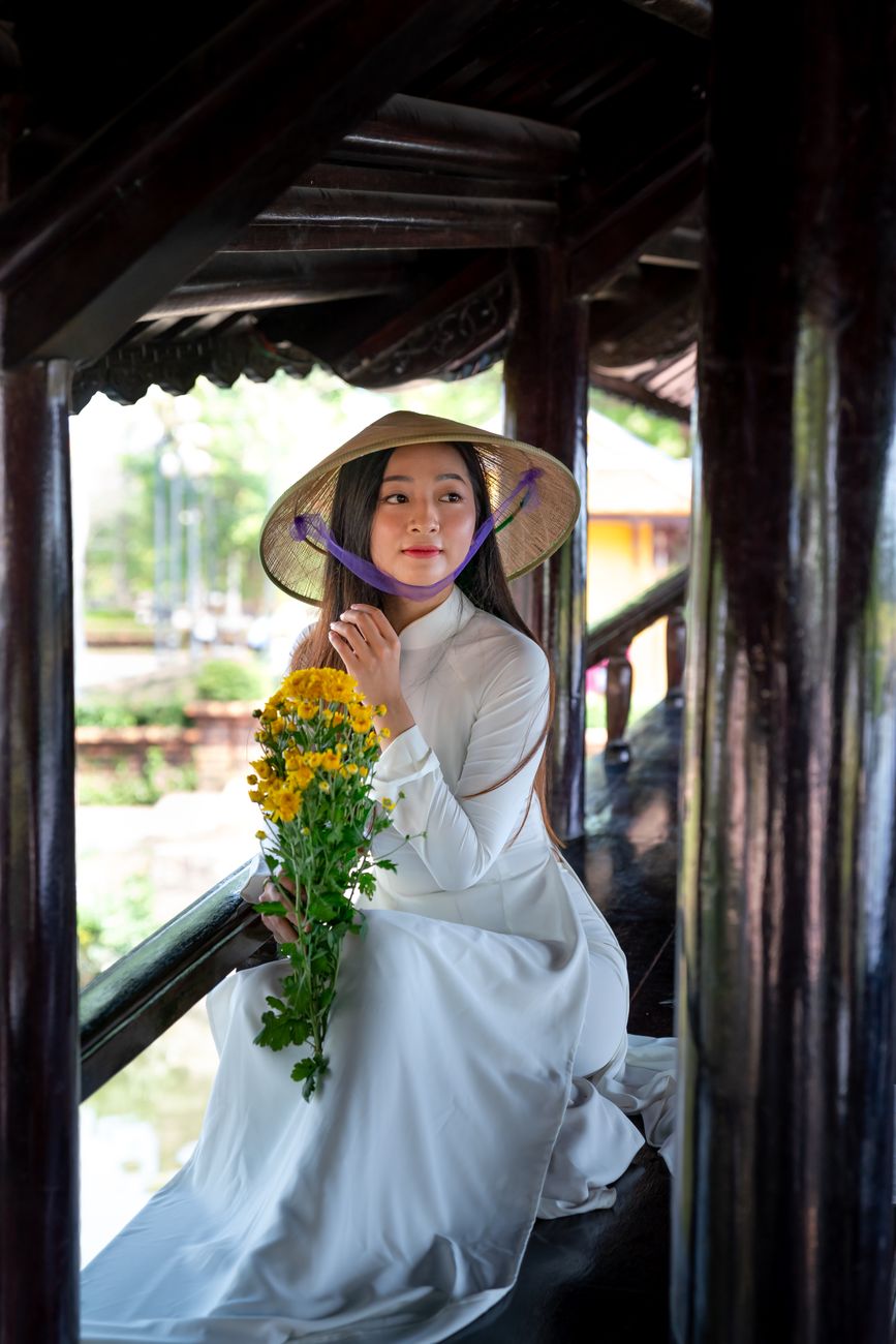 Asian woman in ao dai with conical hat holding yellow flowers indoors.
