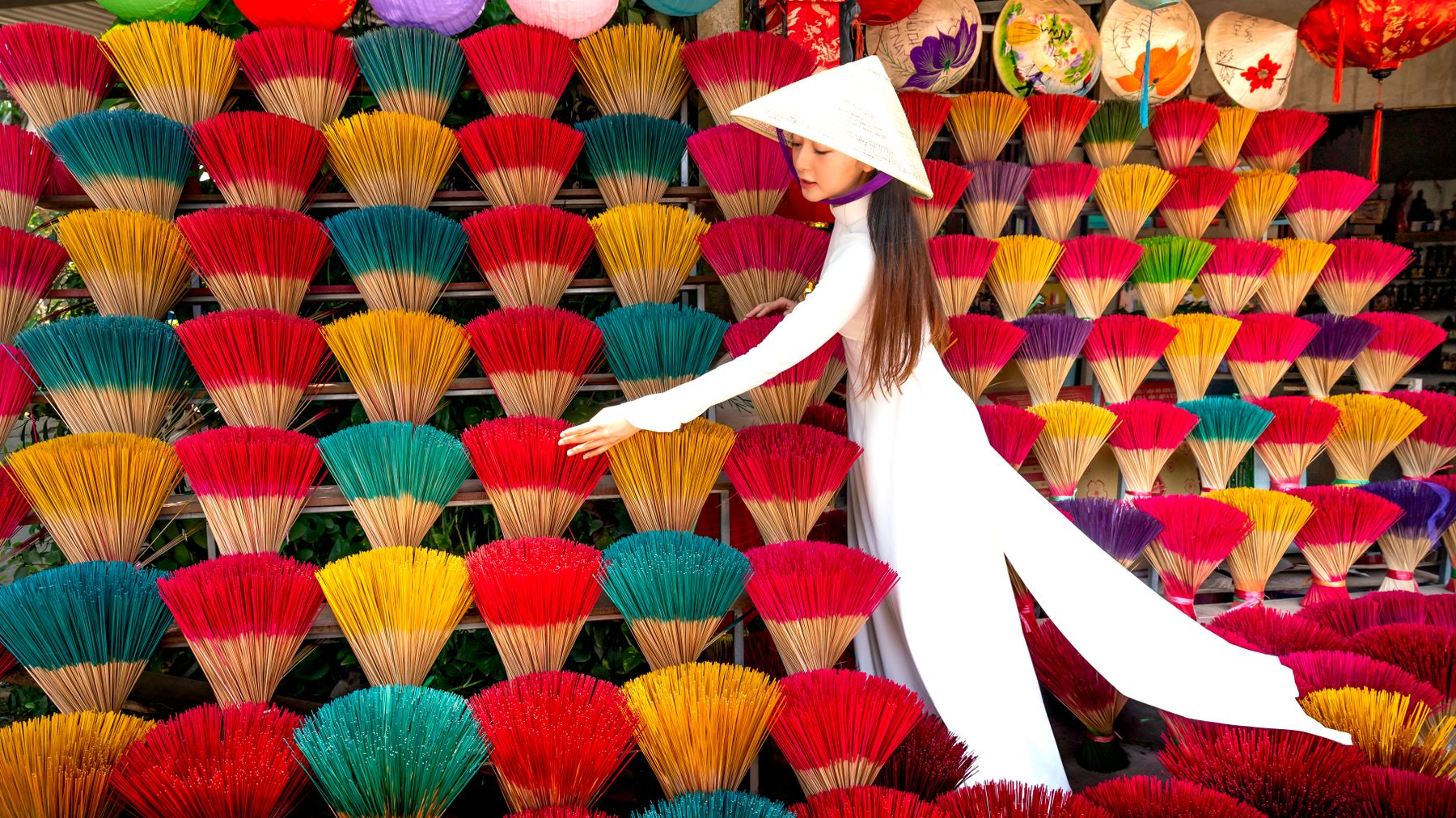 Asian woman in traditional attire arranging colorful incense sticks at a vibrant market stall.