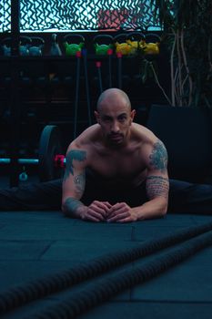 Shirtless man with tattoos stretching on gym floor for workout routine.