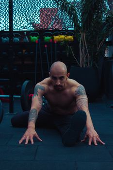 A shirtless man with tattoos stretches on gym floor surrounded by equipment.