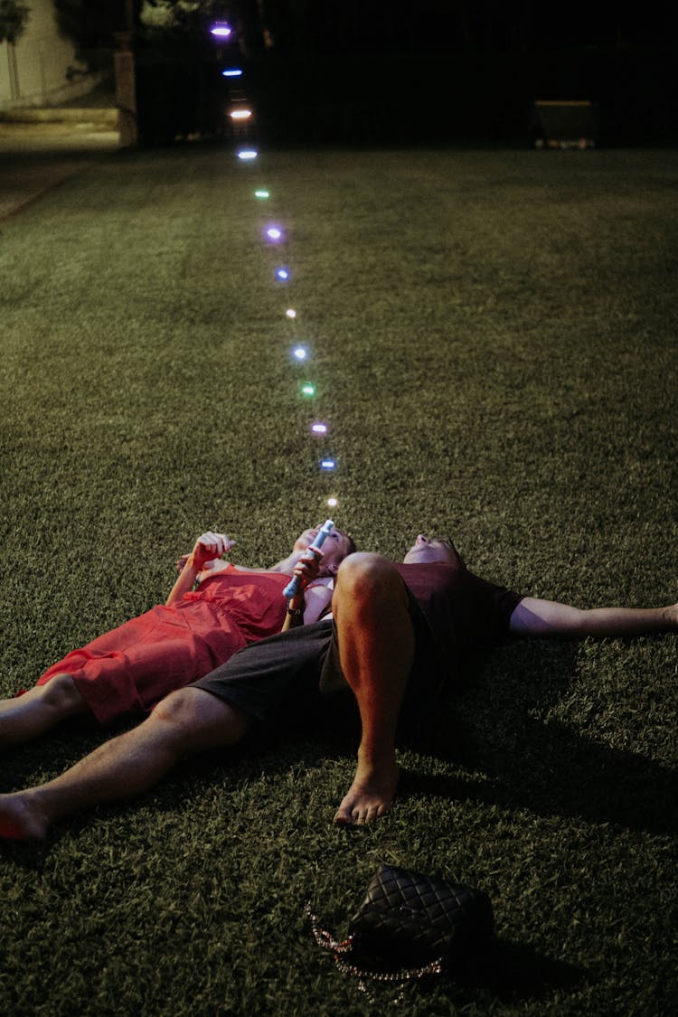 Man And Woman Lying On Green Grass Holding String Lights