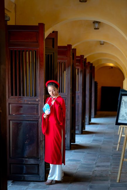 A woman in traditional Vietnamese clothing stands in a hallway holding a fan.