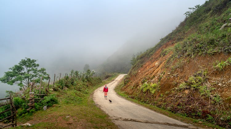 A Woman And A Dog Walking On A Dirt Road Near A Mountain Slope