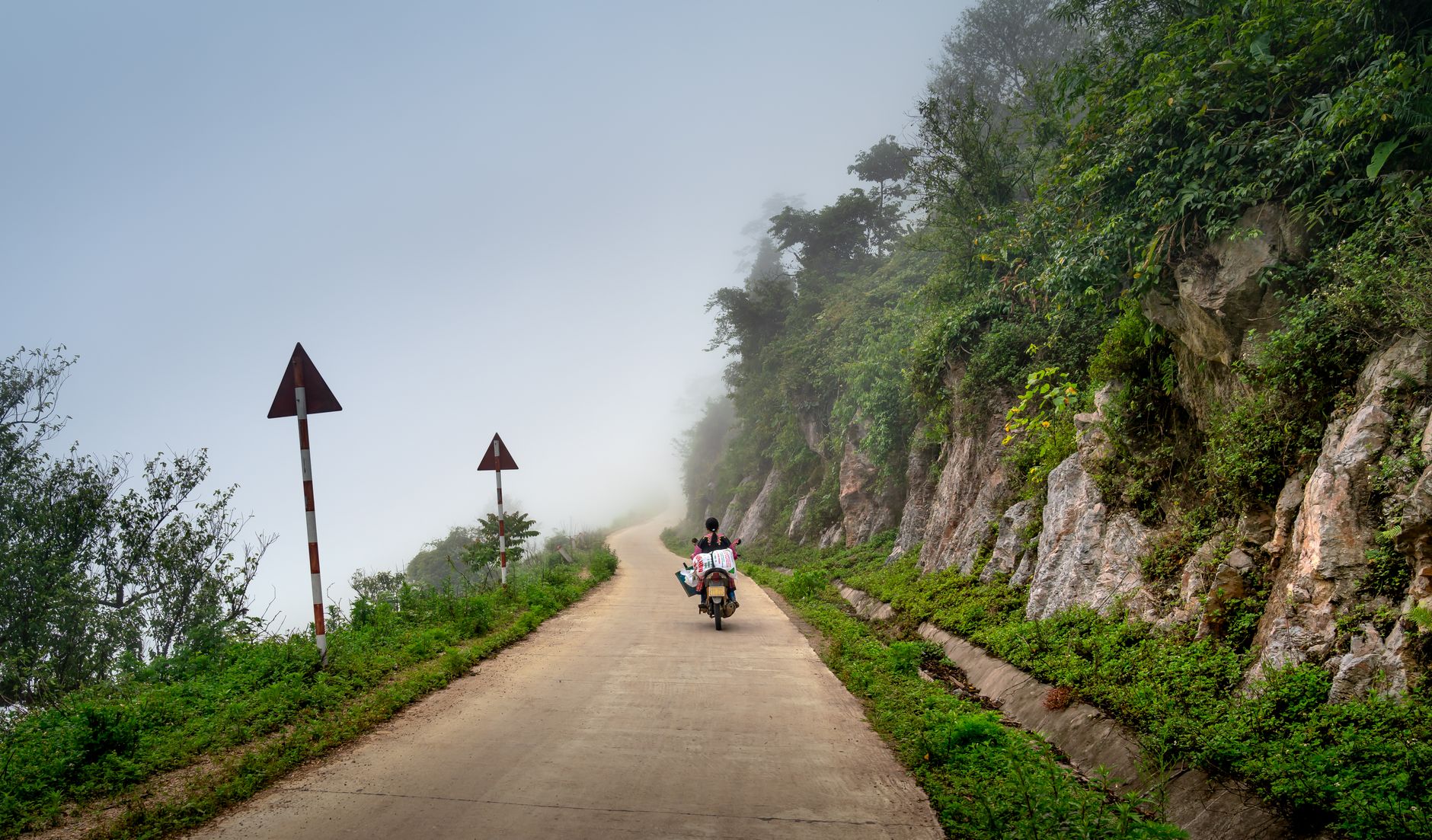 A person rides a motorcycle on a foggy mountain road, surrounded by cliffs and greenery.