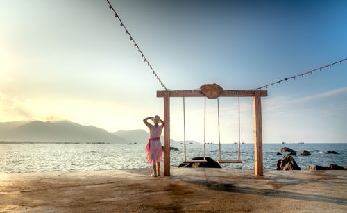 Free A woman stands by a swing on a seaside platform, admiring the ocean horizon at sunset. Stock Photo