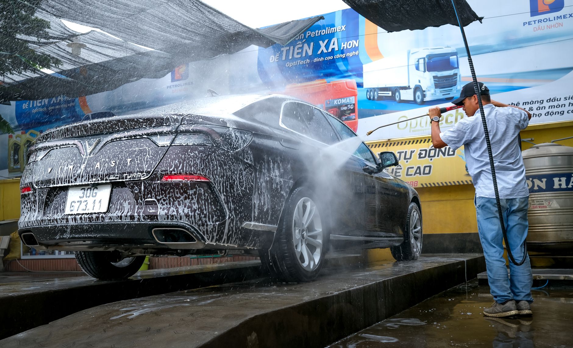 A black car is being cleaned with a power spray by a man at a car wash station outdoors.
