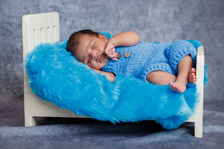 Adorable Infant In Blue Overalls Crochet Clothes Sleeping On Small Bed