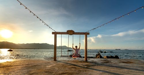 Free Serene scene of a person swinging on the beach at sunset, with ocean and mountains in the background. Stock Photo