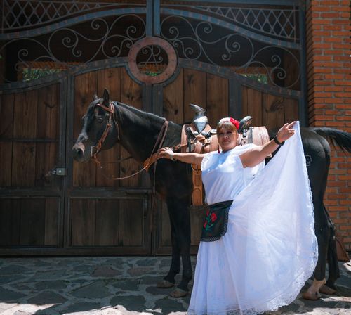 Free A woman in a white dress and traditional attire poses joyfully beside a black horse. Stock Photo