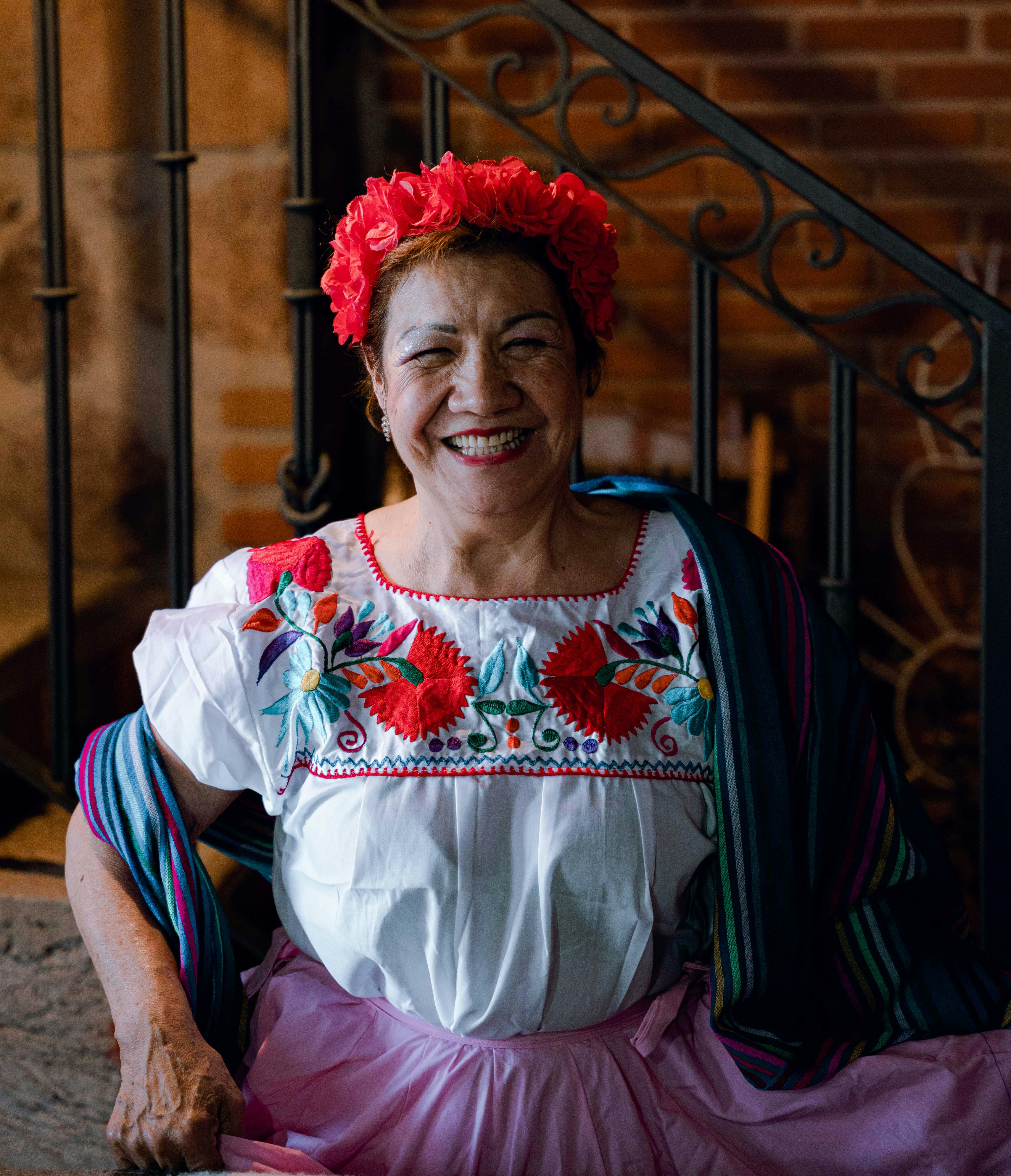 Elderly woman smiling in vibrant traditional attire with floral headpiece indoors.