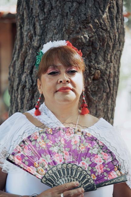 Woman in traditional attire holding a floral fan by a tree outdoors. Bright and colorful setting.