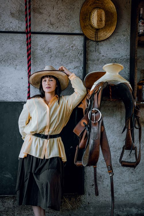 Free Portrait of a woman in a hat leaning against a rustic wall next to a hanging saddle and rope. Stock Photo