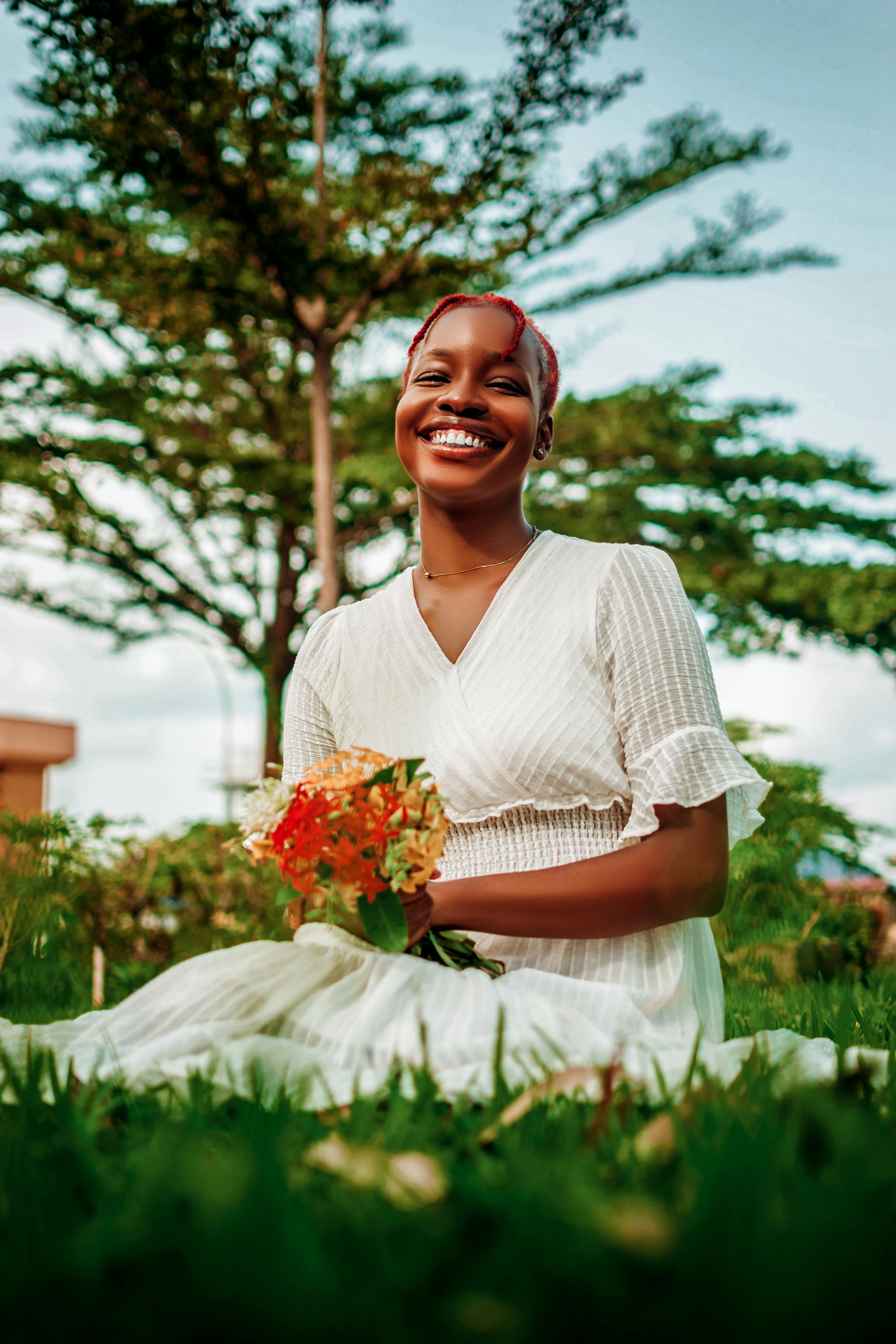 A cheerful woman in a white dress smiles while sitting in a lush green field, holding a bouquet.