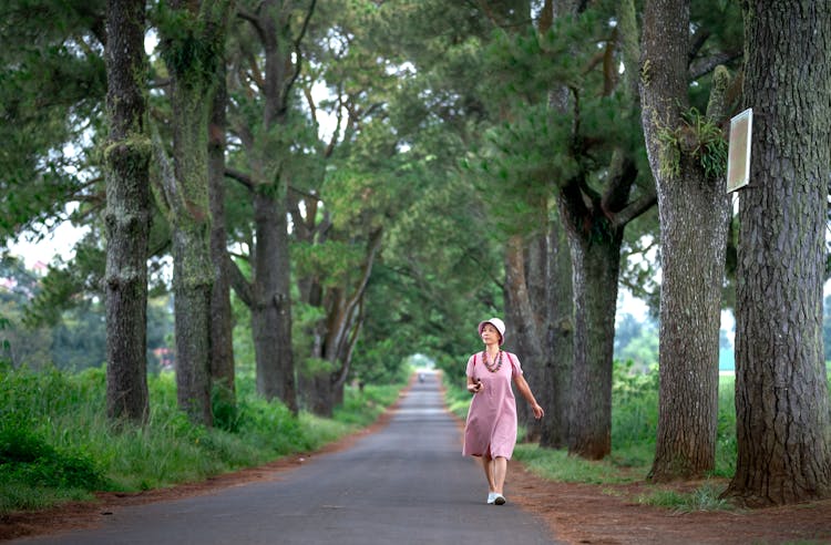 Woman In Beige Dress And Summer Hat Walking Alone On Pathway Between Trees