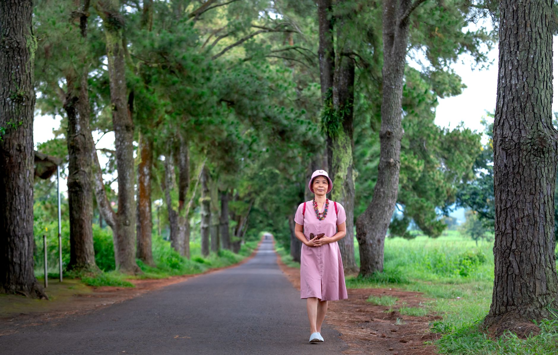 Asian woman walking on road framed by tall pine trees, serene outdoor setting.