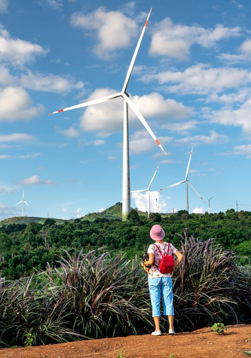 Free Woman with backpack looks at large wind turbines under a blue sky, showcasing renewable energy. Stock Photo