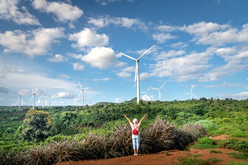 Free A woman joyfully raises her arms in front of wind turbines on a lush green hillside under a blue sky. Stock Photo