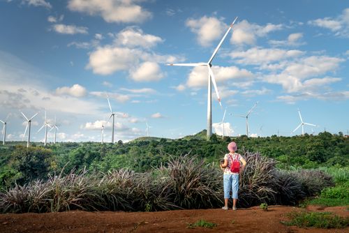 Free A woman stands in a lush countryside, observing wind turbines under a blue sky, symbolizing renewable energy. Stock Photo