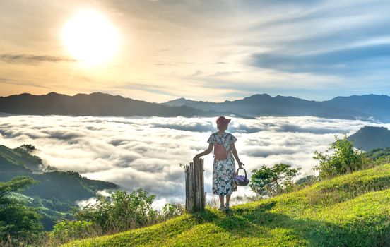 A woman stands on a hillside, overlooking a sunrise above a sea of clouds in the mountains.