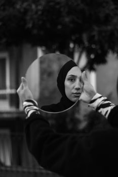 A woman in hijab holds a mirror, reflecting her face, creating a thoughtful scene.