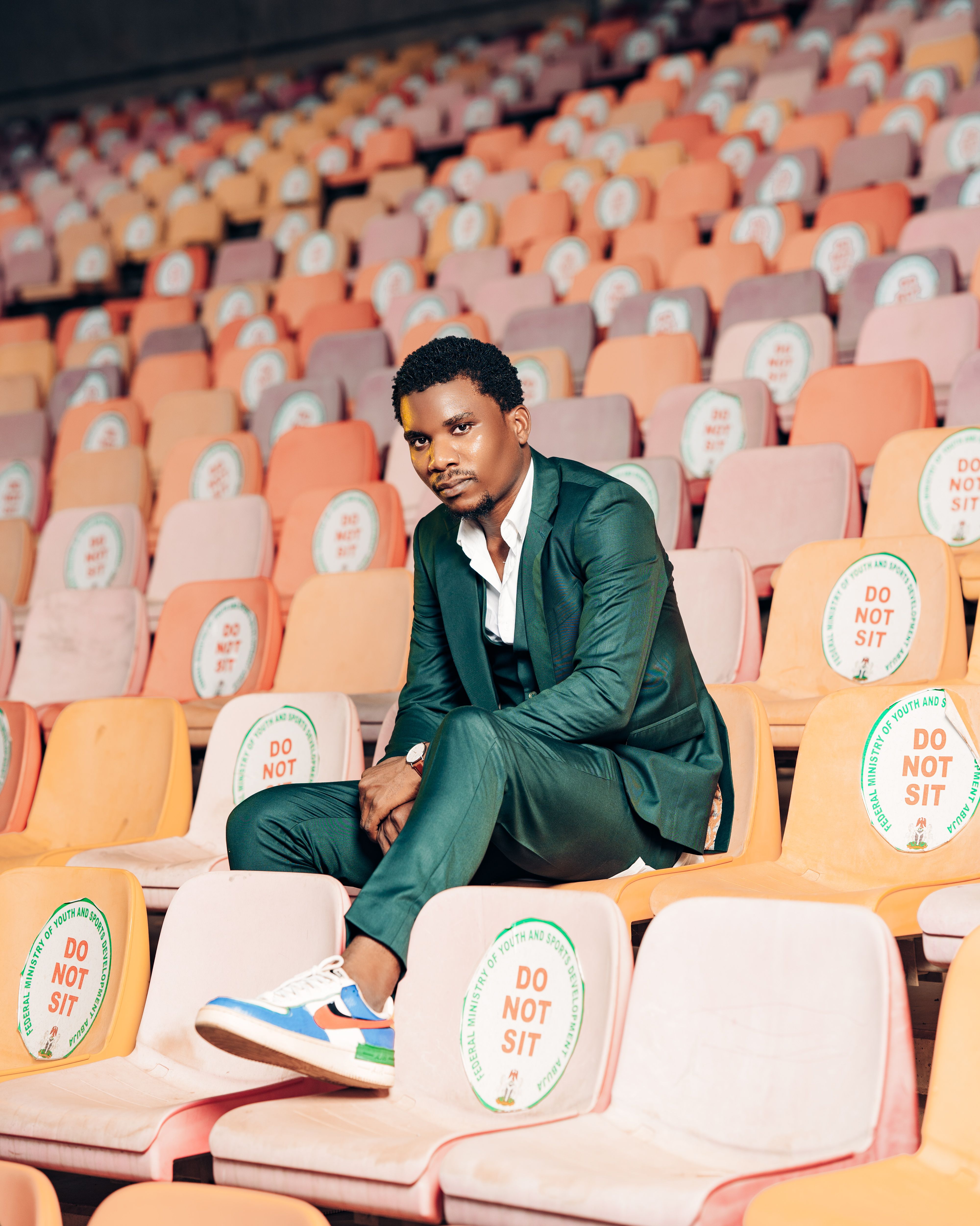Fashionable man in a green suit sitting alone in an Abuja stadium with colorful seats.