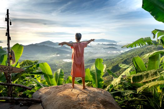 A woman stands on a rock, arms open, overlooking a lush green valley and cityscape.
