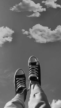 Black and white image of legs in sneakers pointing skyward beneath fluffy clouds.