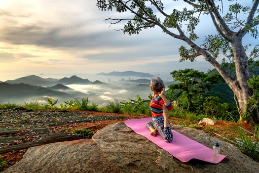 A woman performs yoga on a mountaintop, surrounded by a breathtaking mountain landscape under a serene sky.