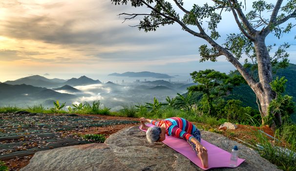 Senior woman doing yoga on a hilltop with a breathtaking mountain view near sunset.
