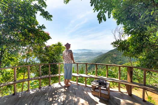 Woman enjoying a panoramic mountain view from a wooden balcony surrounded by lush greenery.