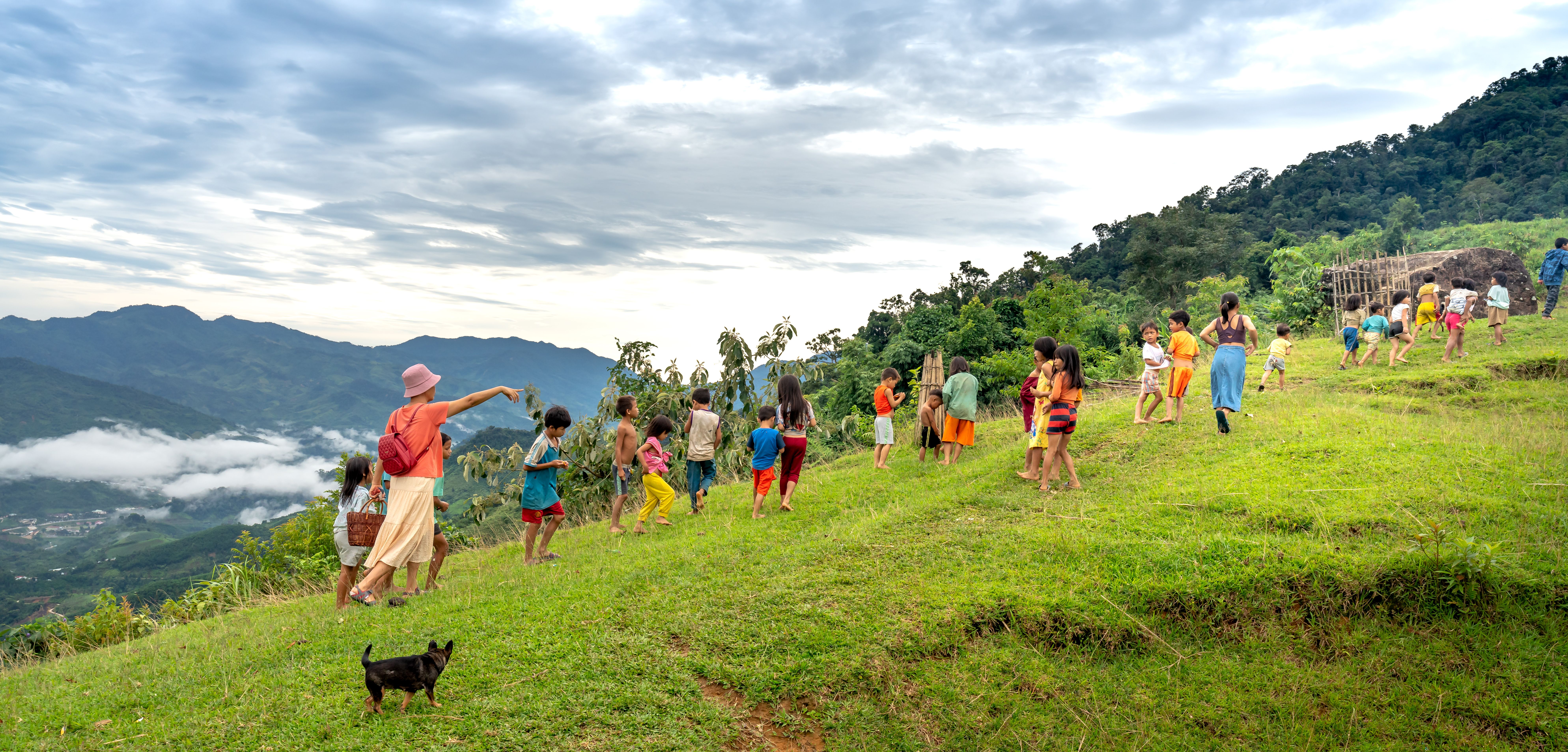 Children and adults enjoying a grassy hillside in a scenic rural Asian landscape.