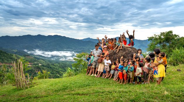 A vibrant group of children joyfully gathered on a hilltop under cloudy skies with scenic mountain views.