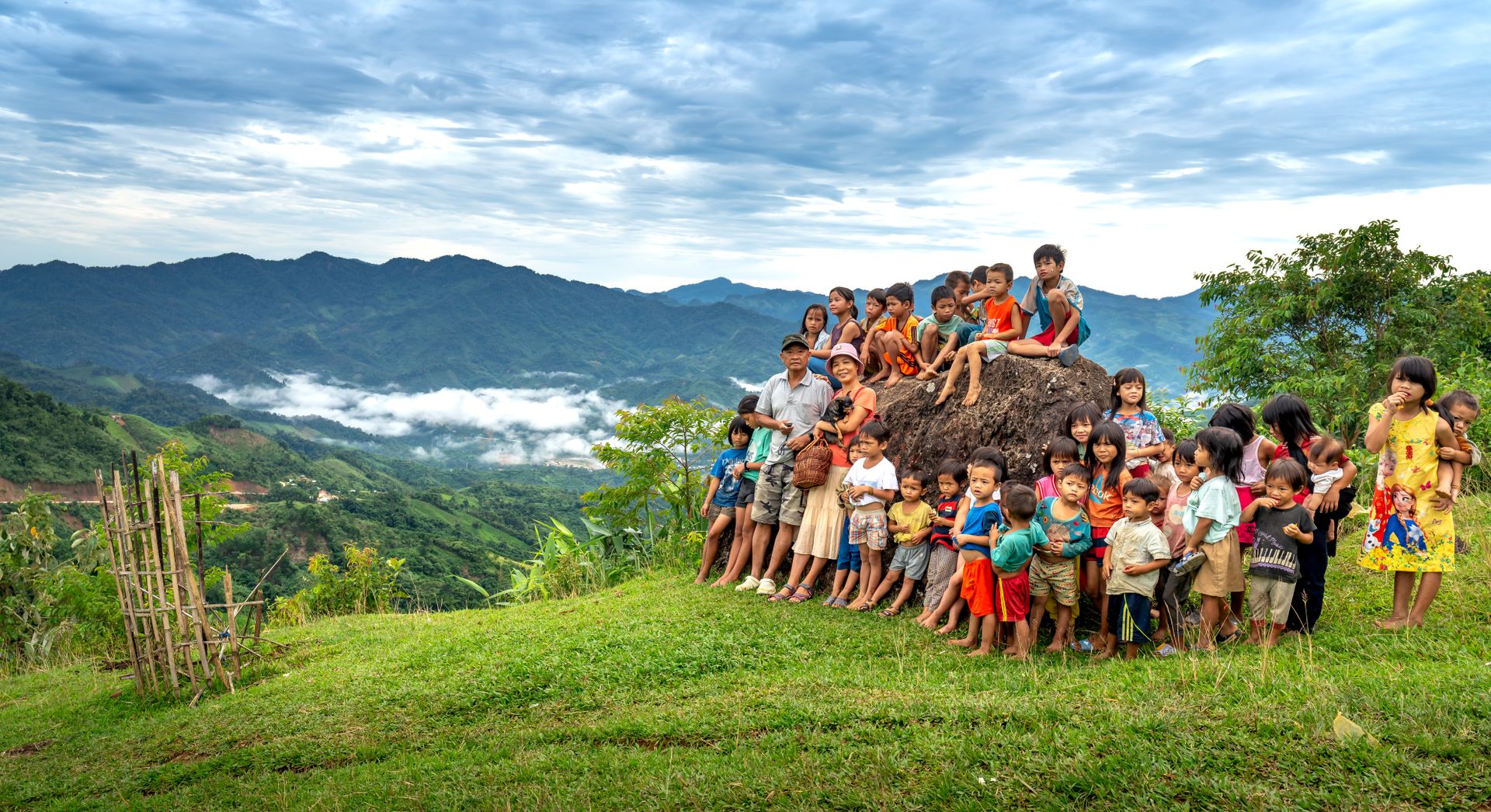 Group of children and adults on a hill overlooking scenic mountains under a cloudy sky.