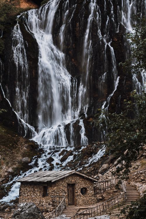 Free A scenic view of a stone house near a cascading waterfall surrounded by lush greenery. Stock Photo