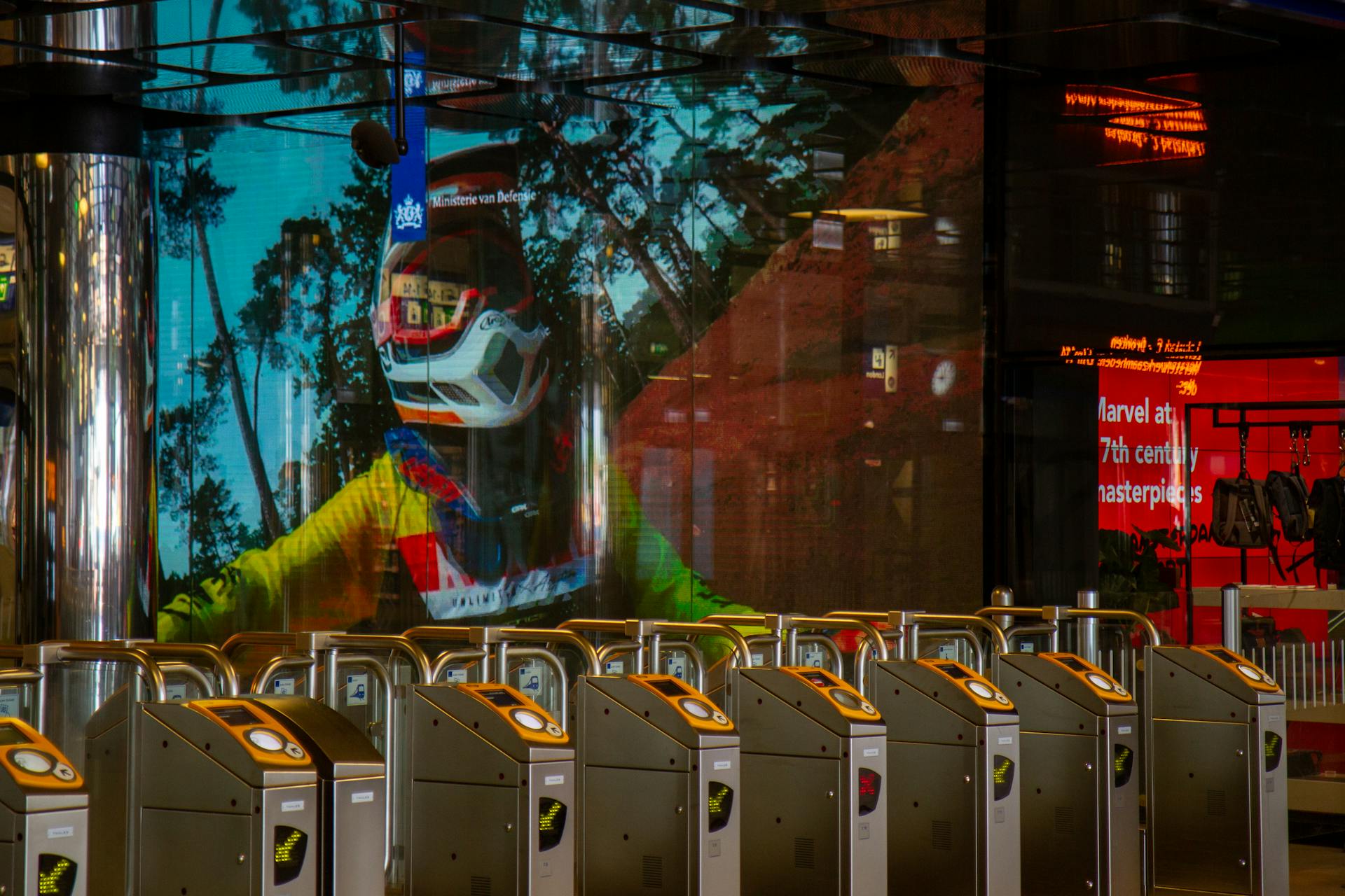 Colorful reflection of a motorcyclist on a large screen at an Amsterdam metro station entrance gate.