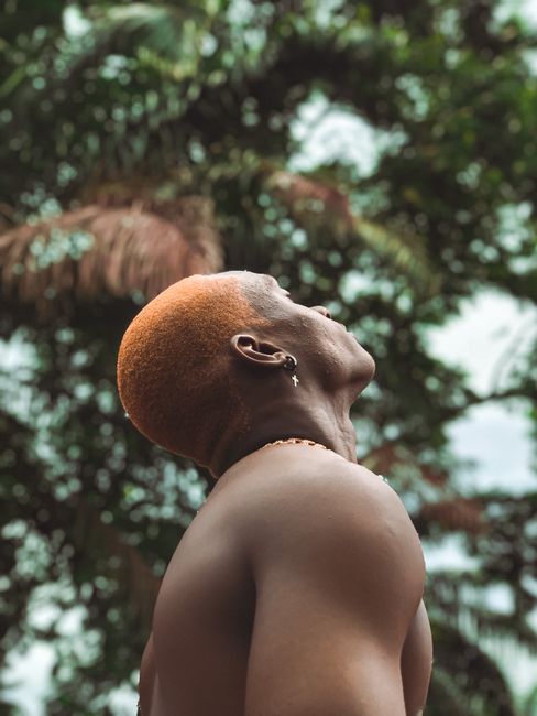 A shirtless man with dyed red hair looking up against a leafy background.