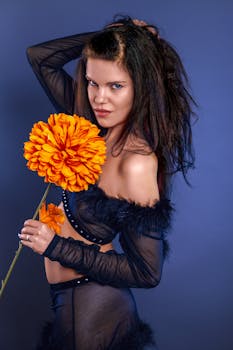 Stylish woman in black outfit posing with bright orange flower against blue backdrop.
