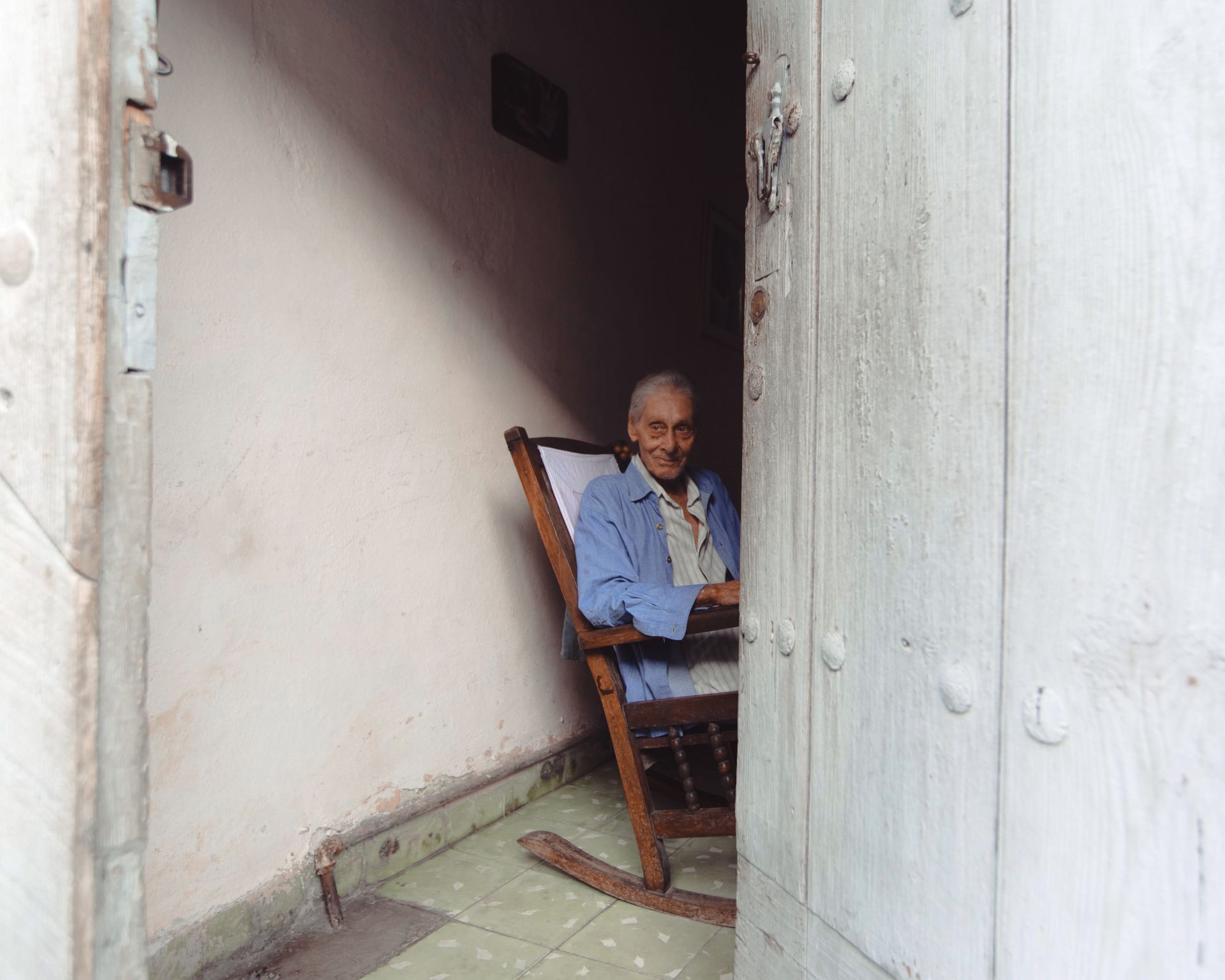 Elderly man in blue shirt sits in wooden rocking chair near open doorway, smiling warmly.