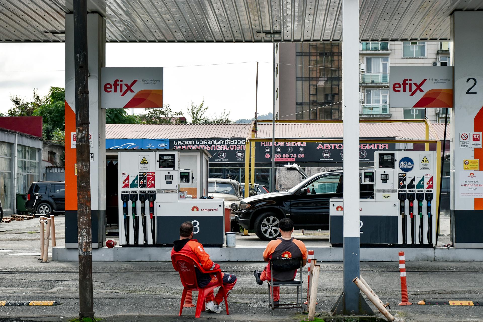 Two men sitting at a gas station with fuel pumps and cityscape background.