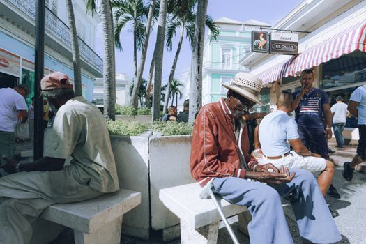 Men sitting on benches in a vibrant street scene with palm trees and a cafe.
