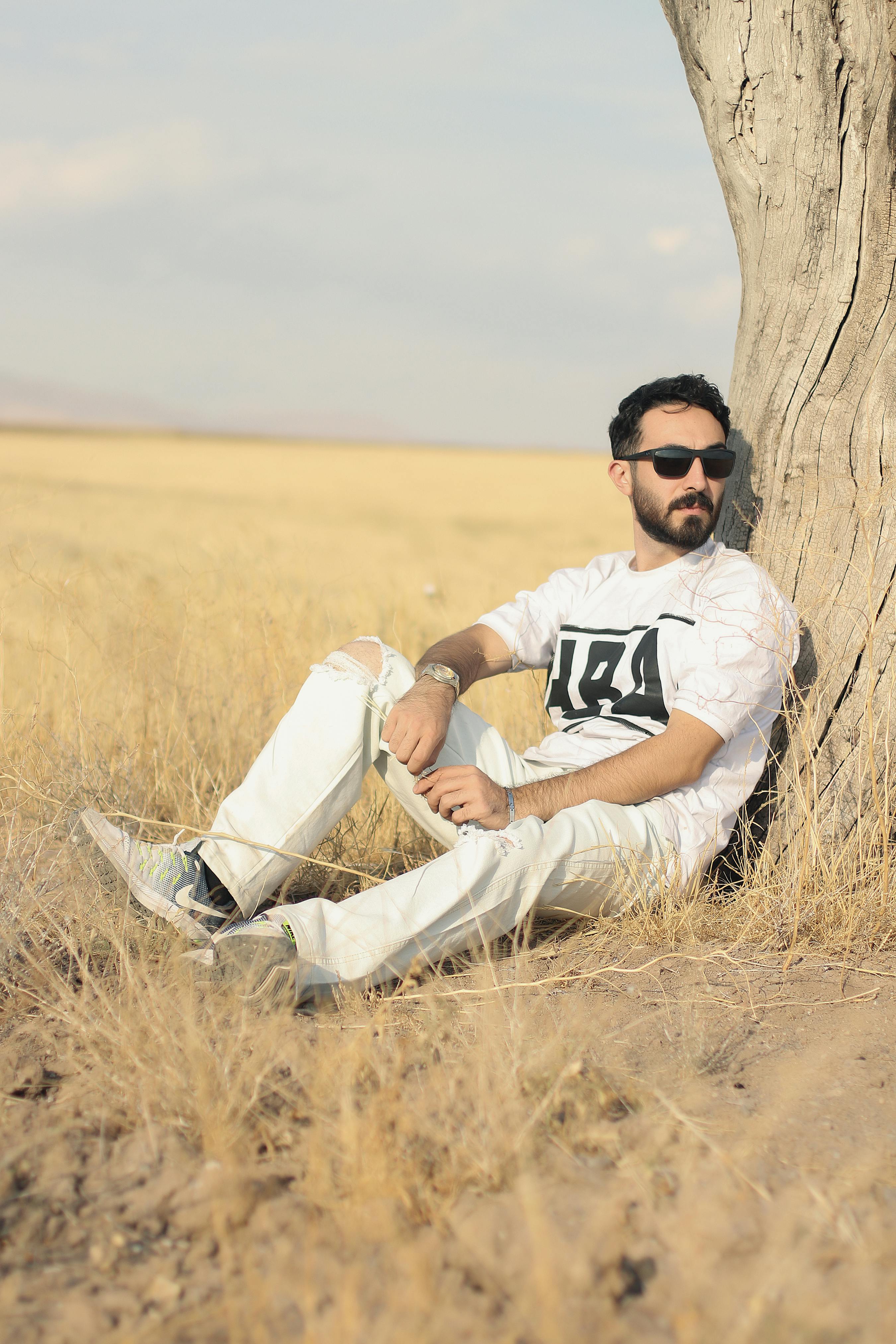 A man with sunglasses relaxes against a tree in an arid, sunny field.