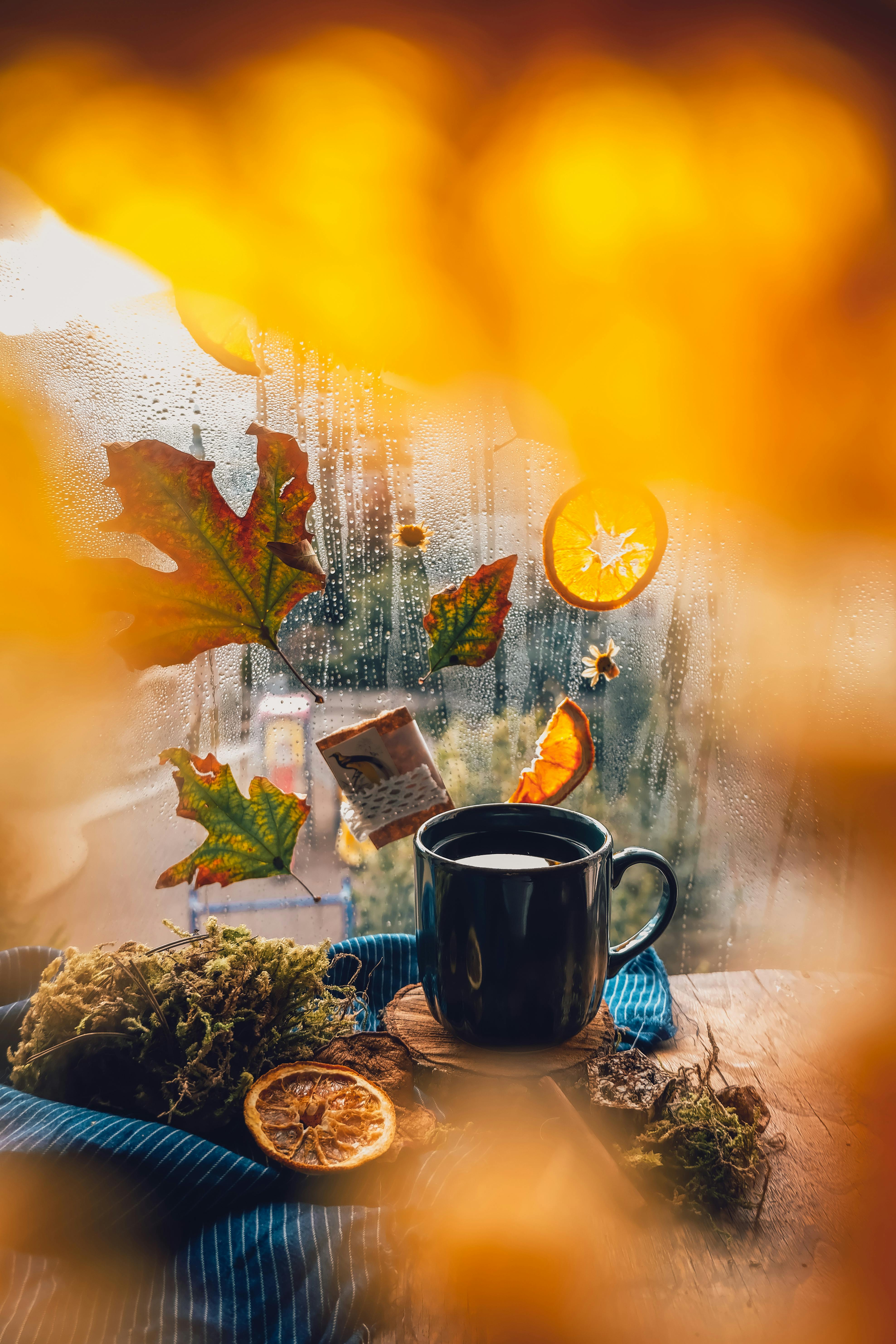 Black ceramic mug on wooden table with autumn leaves and dried orange slices near a glass window.